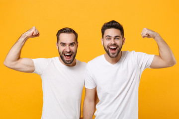 Funny strong young men guys friends in white blank empty t-shirts posing isolated on yellow orange background studio portrait. People lifestyle concept. Mock up copy space. Showing biceps, muscles.