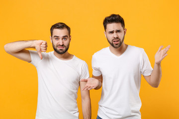 Perplexed young men guys friends in white blank empty t-shirts posing isolated on yellow orange background in studio. People lifestyle concept. Mock up copy space. Showing thumb down, spreading hands.