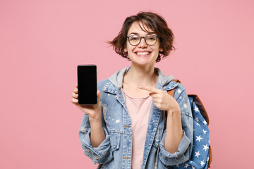 Smiling woman student in denim clothes glasses backpack isolated on pastel pink background. Education in high school university college concept. Point finger on mobile phone with blank empty screen.