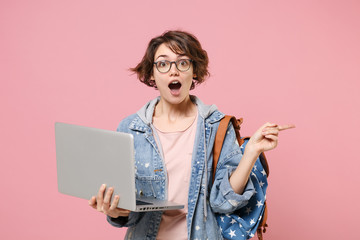 Shocked young woman student in denim clothes glasses backpack isolated on pastel pink background....