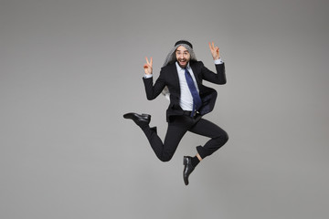 Fototapeta na wymiar Funny young bearded arabian muslim businessman in keffiyeh kafiya ring igal agal classic suit isolated on gray background. Achievement career wealth business concept. Jumping, showing victory sign.
