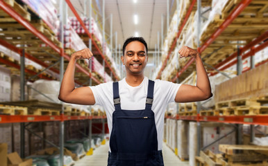 logistic business and people concept - happy smiling indian loader or worker showing his power over warehouse background