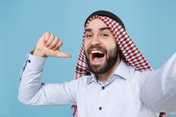 Close up of arabian muslim man in keffiyeh kafiya ring igal agal casual clothes isolated on pastel blue background. People religious concept. Doing selfie shot on mobile phone, point thumb on himself.
