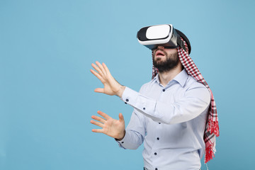 Amazed arabian muslim man in keffiyeh kafiya ring igal agal casual clothes isolated on pastel blue background. People religious lifestyle concept. Watch in headset touch something like push on button.