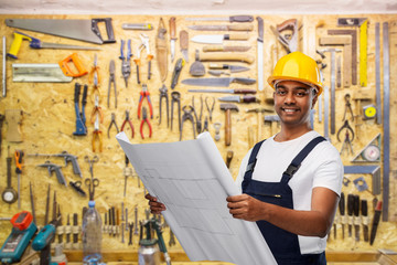 profession, construction and building - happy smiling indian builder in helmet with blueprint over working tools on wall at workshop background