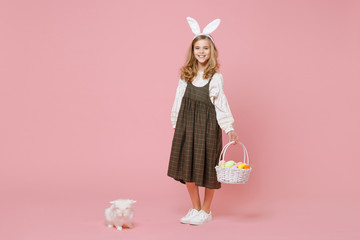 Little pretty blonde kid girl 11-12 years old in light spring dress hold fluffy white bunny rabbit, wicker basket with eggs isolated on pastel pink background. Childhood lifestyle Happy Easter concept