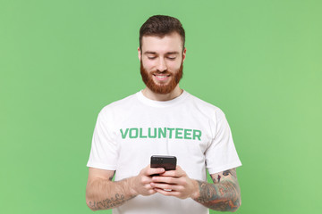 Smiling young bearded man in white volunteer t-shirt isolated on pastel green background. Voluntary...