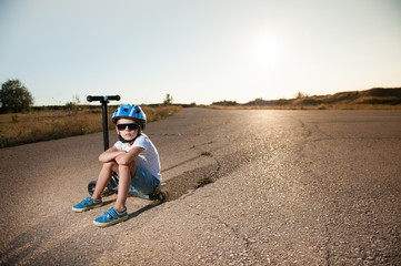 tired active little caucasian boy in sport blue helmet and sneakers sitting on his black scooter on hot summer asphalt road on sunset with copy space