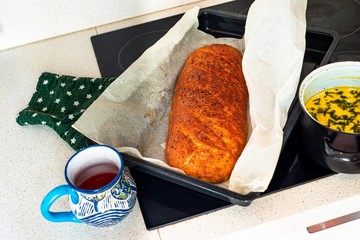 Freshly baked bread in baking tray, pot with soup, cup of tea on kitchen board.