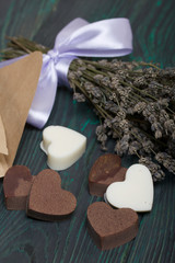 An open white paper bag on brushed pine boards. Chocolate hearts are scattered from it. From white and black chocolate. Near a bouquet of lavender.