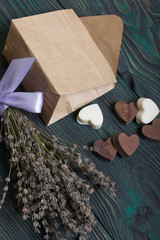 An open white paper bag on brushed pine boards. Chocolate hearts are scattered from it. From white and black chocolate. Near a bouquet of lavender.