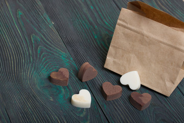 An open white paper bag on brushed pine boards. Chocolate hearts are scattered from it. From white and black chocolate.