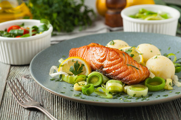 Portion of fried salmon, served with mashed potatoes and cooked leek. Front view. Gray plate, wooden background.