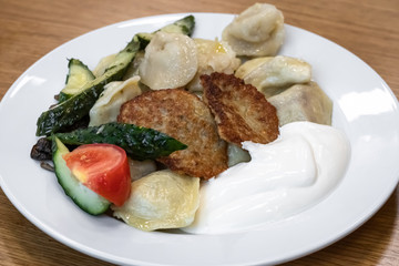 A heap dumplings and fried potato pancakes with sour cream on white plate on wooden background. Traditional Russian dish. Ukrainian cuisine.