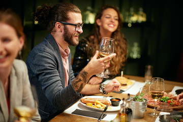 Handsome caucasian bearded hipster man with eyeglasses smiling and holding glass of wine while sitting with his friends in restaurant.