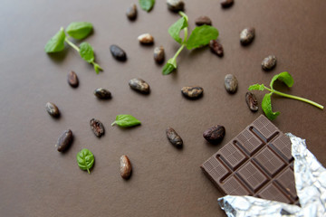 sweets, confectionery and food concept - dark chocolate bar in foil wrapper with peppermint and cocoa beans on brown background