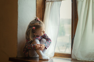beautiful little girl in a simple dress sits on a wooden window of an old village house and admires...