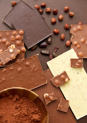 sweets, confectionery and food concept - milk, dark and white chocolate bars with hazelnuts, cocoa beans and powder on brown background