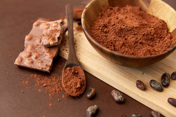 sweets, confectionery and culinary concept - chocolate with hazelnuts, cocoa beans and powder in wooden bowl with spoon on brown background