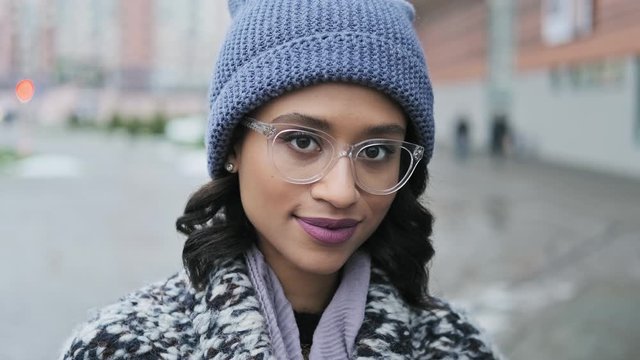 Attractive young black woman in glasses look at camera and smiling. Portrait of beautiful trendy african american woman in hat and coat outdoors. Close-up of fashion model in 4K, UHD