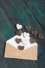An open white paper envelope on brushed pine boards. Chocolate hearts are scattered from it. From white and black chocolate.