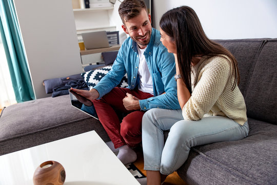 Smiling couple sitting on sofa at home spending time together while using tablet.