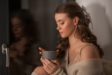 winter, comfort and people concept - young woman in pullover sitting at window with coffee or tea cup at home
