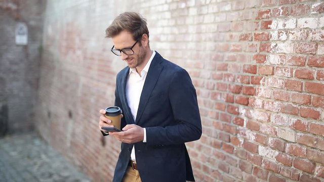young smiling man wearing glasses is reading on his phone and starts to walk  while drinking coffee