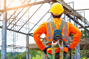 Construction worker wearing safety harness and excavator wearing orange reflective vest standing in...