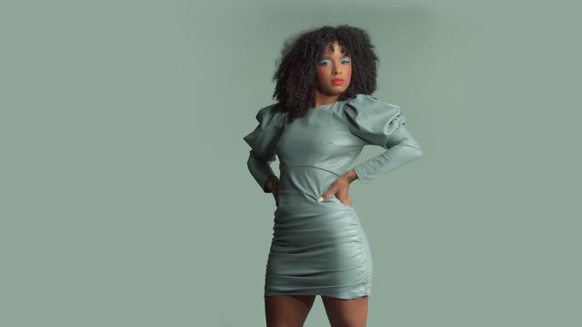 Mixed race woman with huge afro curly hair, wears leather dress on the same background color moves and poses to the camera with hands on her waist