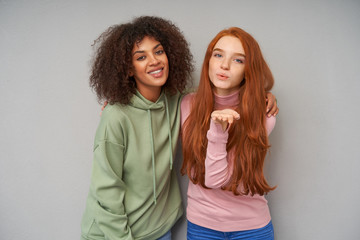 Indoor photo of cheerful young lovely girlfriends hugging each other while posing over grey background in comfortable casual clothes, looking positively at camera