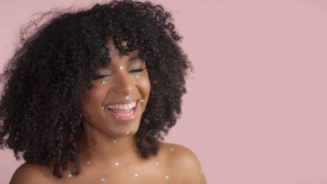 Mixed race black woman with curly hair covered by crystal makeup on pink background in studio Happy smiling laughing to the camera