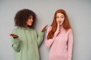 Displeased young pretty curly dark skinned brunette female looking with dissapointed face at her redhead girlfriend and raisingmemotionally hands, isolated over grey background