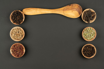 Six small wooden bowls filled with different types of peppercorns and a wooden cooking spoon made of olive wood lie on a black background with copy space in the center