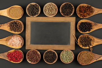 Six small wooden bowls and eight cooking spoons made of olive wood filled with various spices and an old black slate board with copy space lie on a black background
