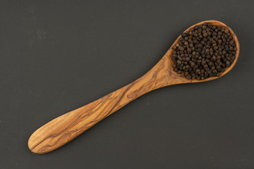 A cooking spoon made of olive wood with black peppercorns on a black background with copy space