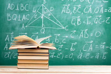 Education and reading concept - group of books on the wooden table, green blackboard with formulas on the background. Teachers desk