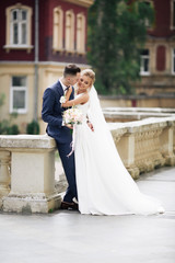Naklejka premium Wedding photo shooting. Bride and bridegroom walking in the city. Married couple embracing and looking at each other. Holding bouquet. Outdoor, full body