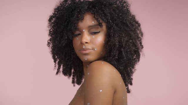 Mixed race black woman with curly hair covered by crystal makeup on pink background in studio turns to the camera from the profile position