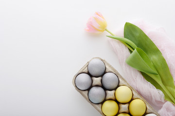 Easter eggs in pastel color in cardboard box, pink-yellow Tulip on pink textile towel on white background