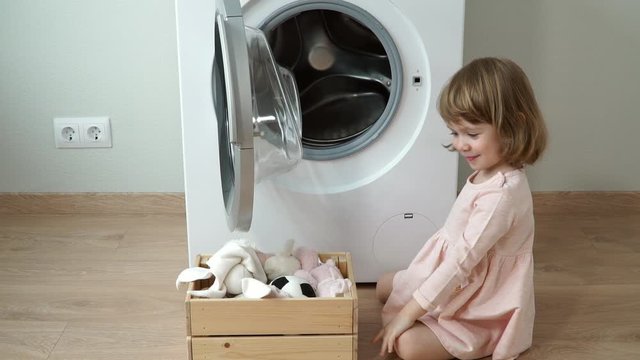 Portrait of little smiling cute 3 year old girl sits on laminate floor, opens hatch and puts in washing machine soft toys for washing