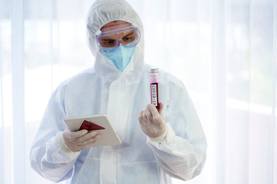 Doctor in personal protective suit looking at the Coronavirus COVID 19 blood sample tube and holding a tablet computer.