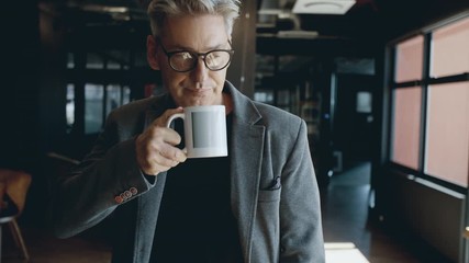 Happy mature businessman wearing spectacles drinking coffee in office. Successful businessman smiling in a creative office having a coffee break. - Powered by Adobe