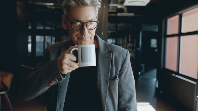 Happy mature businessman wearing spectacles drinking coffee in office. Successful businessman smiling in a creative office having a coffee break.