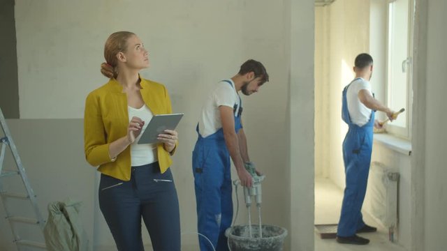 Woman walks with tablet, repairmen work in the flat