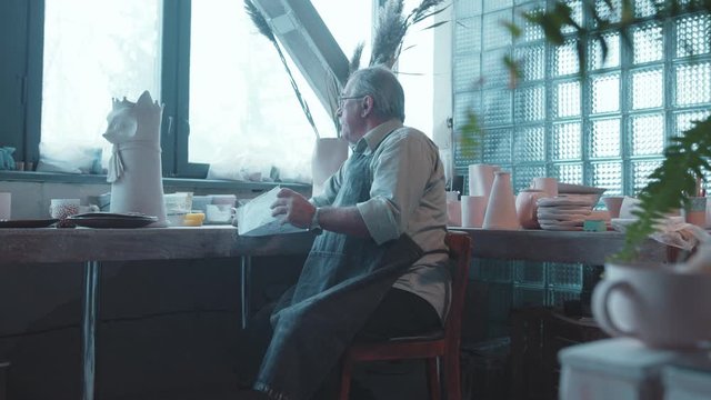 Mature caucasian potter in muddy apron in his workshop looking at old child painting recovering family memories sitting happy. Pottery.