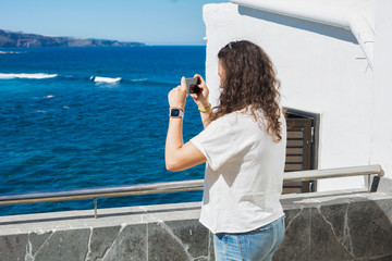 Woman taking photos with her smartphone to a summer landscape with sea and white houses lit by the sun.