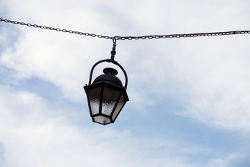 Fototapeta na wymiar Closeup of vintage street light suspended on metallic chain in the street on cloudy sky background