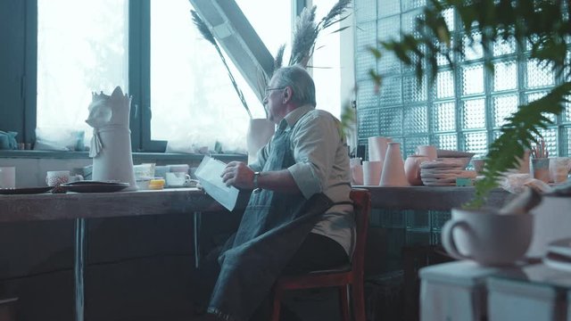 Professional old bearded potter in muddy apron looks at kis art sketch thinking of ideas creating and working in pottery workshop.