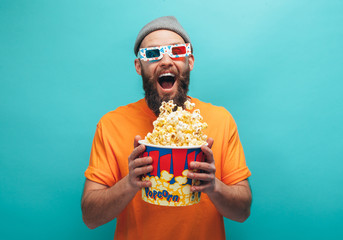 Handsome emotional young guy in orange t-shirt eat popcorn wearing 3d glasses watch film, isolated on blue background.